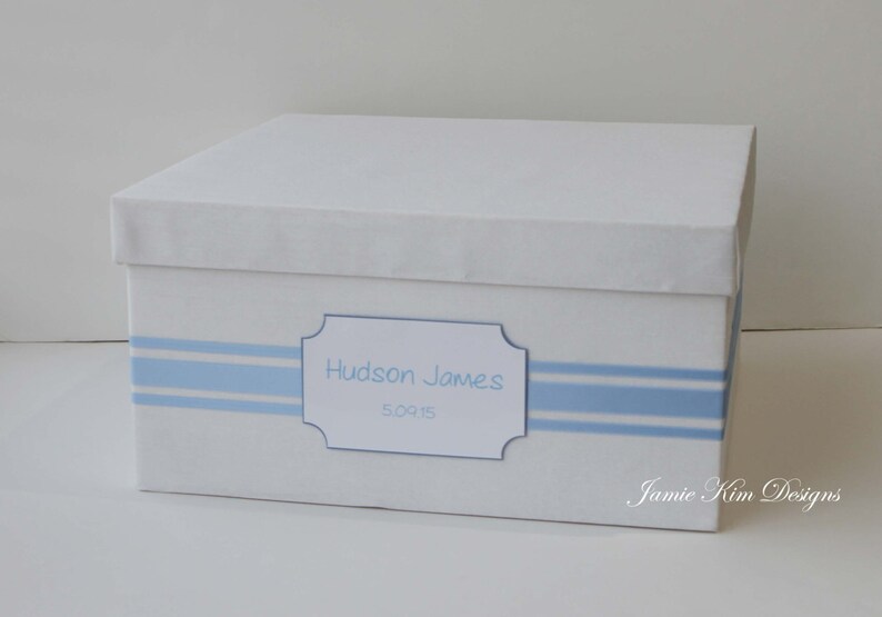 May include: White keepsake box with blue stripes and a label that reads "Hudson James 5.09.15". The box is decorated with a blue ribbon and the words "Jamie Kim Designs" are printed on the bottom right corner.