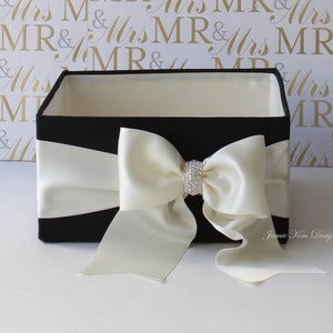 Pode incluir: Caixa de presente preta com um laço de cetim branco e um enfeite de strass. A caixa está aberta e vazia. O texto "Janie Kim Designs" é visível no canto inferior direito.