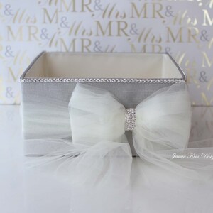 May include: A gray square box with a white tulle bow and rhinestones. The box is decorated with a silver trim. The box is sitting on a white surface. Jamie Kim Designs is printed on the tulle.