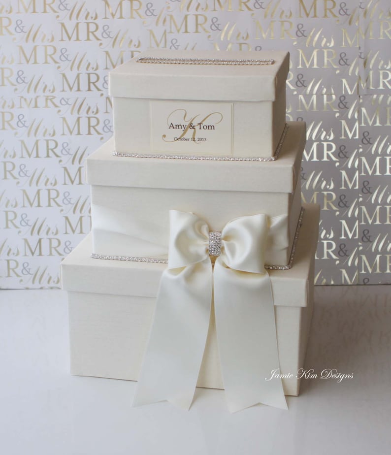 May include: A three-tiered white gift box with a satin ribbon and a rhinestone embellishment. The top box has a personalized label with the names "Amy & Tom" and the date "October 12, 2013".
