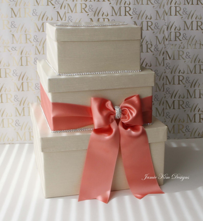 Puede incluir: Tres cajas de regalo de color marfil apiladas una encima de la otra, atadas con una cinta de sat&eacute;n coral y un gran lazo. Las cajas est&aacute;n decoradas con una fila de diamantes de imitaci&oacute;n alrededor del borde superior.