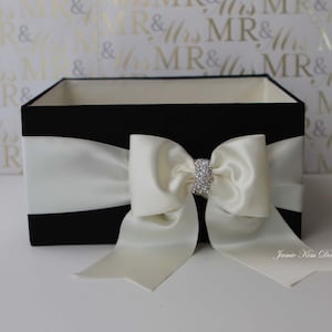 May include: Black velvet gift box with a white satin bow and a rhinestone embellishment. The box is sitting on a white surface with a gold and white pattern.