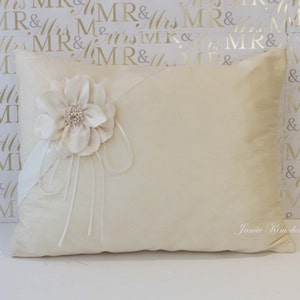 Könnte beinhalten: Ein weißes Satin-Kissen mit einer großen weißen Satin-Blume und einem weißen Satin-Band. Das Kissen liegt auf einem weißen Hintergrund mit einem goldenen Muster aus wiederholtem "MR & Mrs".