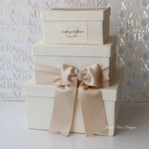 May include: Three white gift boxes stacked on top of each other. The top box has a gold ribbon and a label that says "Zachary & Jillian" and "May 17, 2014". The middle box has a large beige satin bow. The bottom box has a smaller beige satin bow. The boxes are on a white background with a gold pattern.