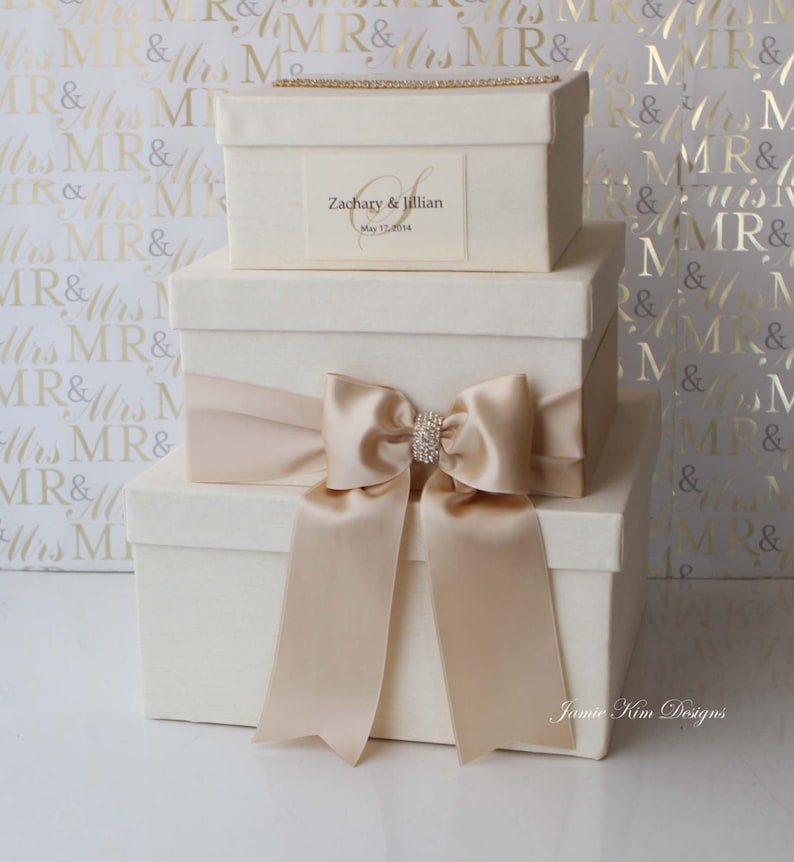 May include: Three white gift boxes stacked on top of each other. The top box has a gold ribbon and a label that says "Zachary & Jillian" and "May 17, 2014". The middle box has a large beige satin bow. The bottom box has a smaller beige satin bow. The boxes are on a white background with a gold pattern.