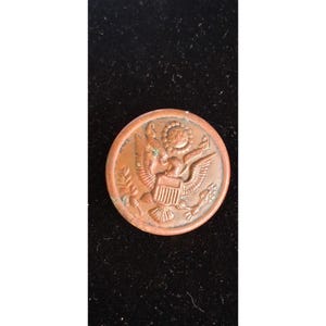 Może przedstawiać: Miedziany guzik z Wielką Pieczęcią Stanów Zjednoczonych. Projekt zawiera orła z rozpostartymi skrzydłami, tarczę i gałązki oliwne. Guzik ma lekko postarzały wygląd.