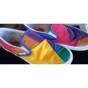 May include: Colorful slip-on sneakers with a patchwork design. The shoes feature panels of red, purple, blue, yellow, and green, with glitter accents. The soles are white, and the interior is white with the brand name printed in red.