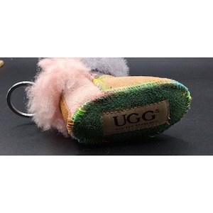 May include: A miniature UGG boot keychain featuring a mix of materials and colors. The keychain has a pink, tan, and green design with a metal ring. The UGG brand logo is visible on the green sole.