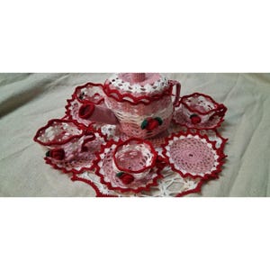 May include: A crocheted tea set in pink and white with red trim. The set includes a teapot, cups, saucers, and a doily. Each piece is adorned with a small red rose. The tea set is handmade.