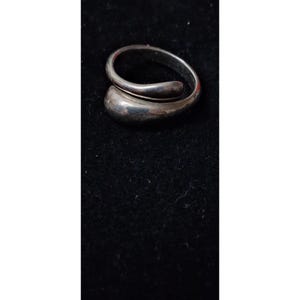 May include: A silver-toned bypass ring featuring a smooth, polished finish. The band curves around to form an open, overlapping design with a rounded, teardrop-shaped end that tapers into a thinner, continuous metal loop.