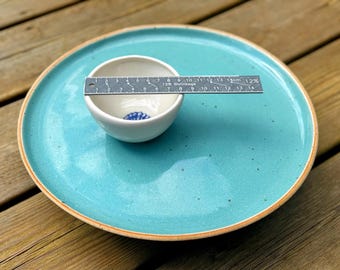 May include: A small, white ceramic bowl with a blue design inside sits on a large, turquoise plate. A ruler is placed across the bowl, showing measurements in inches. The plate has a brown rim and is on a wooden surface.