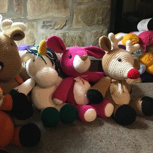 May include: A collection of handmade crochet stuffed animals, including a reindeer, a unicorn, and a pink bunny. The toys have various colors, including white, brown, pink, and green. The toys are sitting on a gray surface.