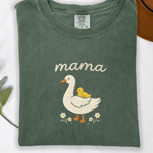 May include: A forest green crewneck t-shirt featuring an embroidered design of a white goose with a small yellow duckling on its back. The word mama is embroidered in white above the goose, with small white flowers at the bottom of the graphic.