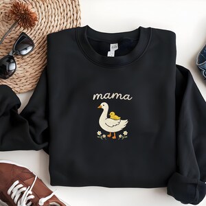 May include: A black crewneck sweatshirt featuring an embroidered graphic of a white goose with a small yellow duckling on its back. The word "mama" is stitched in a light tan script above the goose, surrounded by small white flowers.