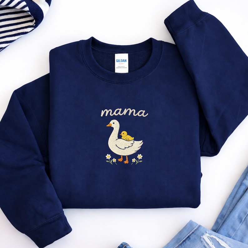 May include: A navy blue crewneck sweatshirt featuring an embroidered design of a white goose with a small yellow duckling on its back. Above the goose, the word mama is embroidered in white script. Small white flowers are at the base of the goose.