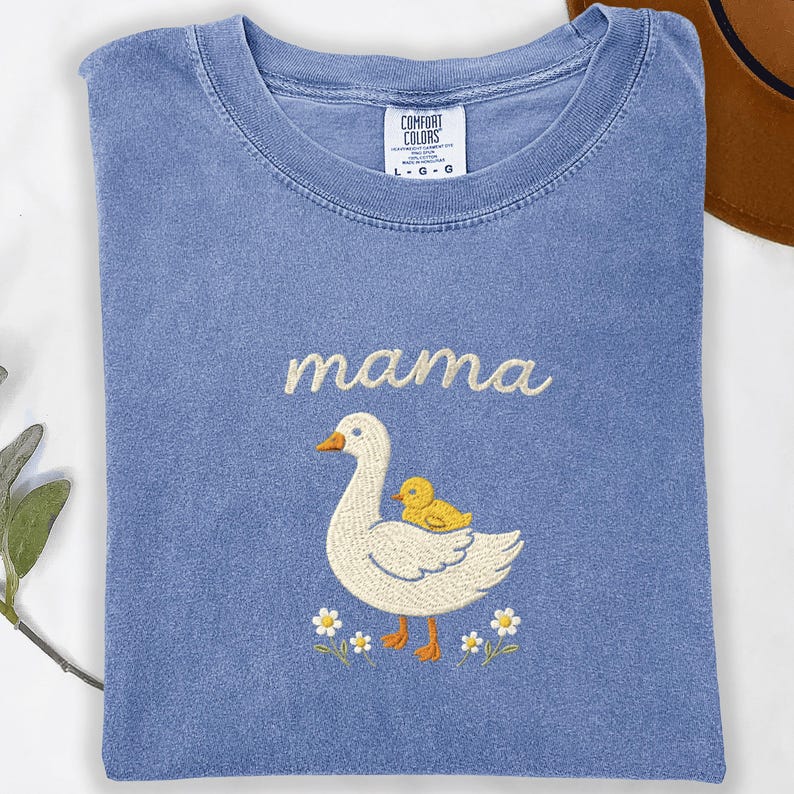 May include: A blue crew neck t-shirt featuring an embroidered design of a white goose with a yellow duckling on its back. Above the goose, the word "mama" is embroidered in white script. The shirt has a Comfort Colors label at the neck.