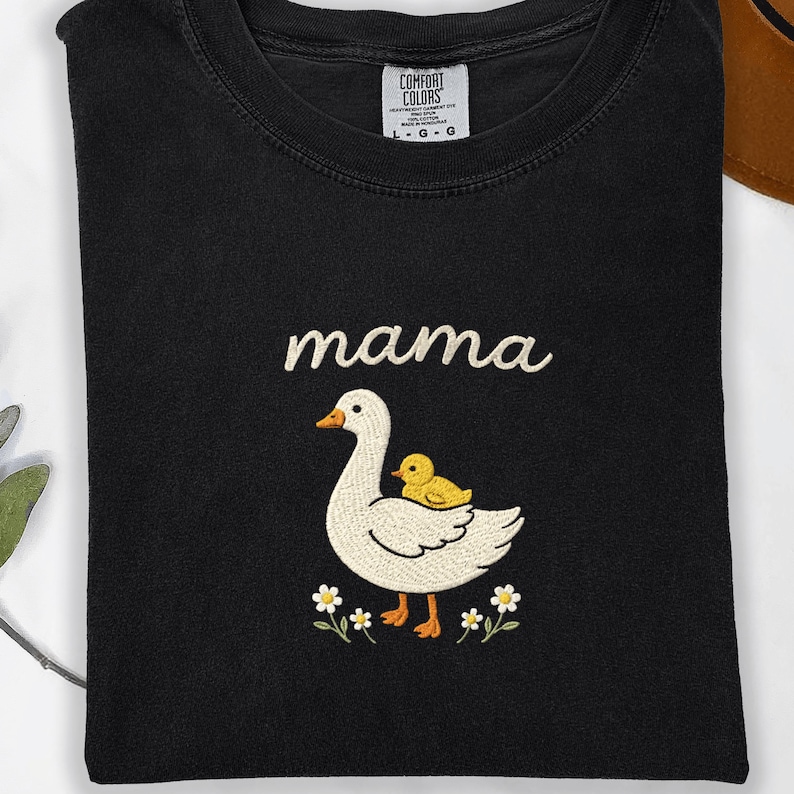 May include: A black crewneck t-shirt featuring an embroidered design of a white goose with an orange beak and feet, carrying a small yellow duckling on its back. The word mama is embroidered in white above the goose, with small white flowers below.