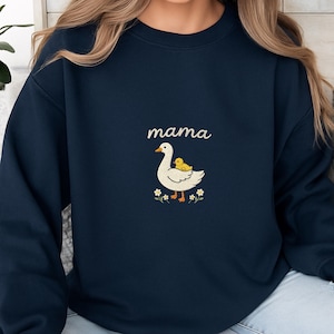 May include: A navy blue crewneck sweatshirt featuring a central graphic of a white goose with an orange beak and feet, carrying a small yellow duckling on its back. Above the goose is the word mama in white script, surrounded by small white flowers.