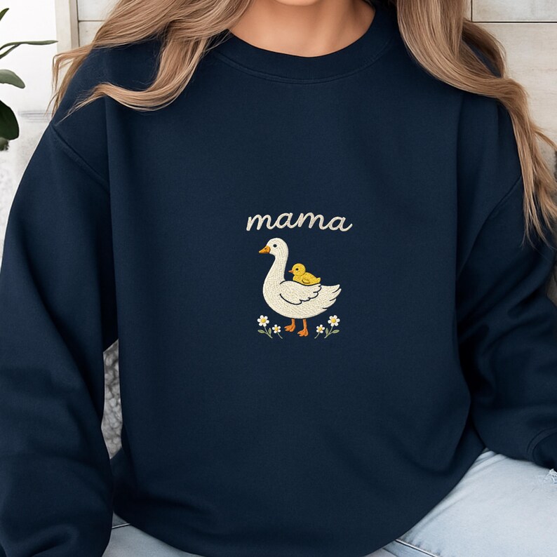 May include: A navy blue crewneck sweatshirt featuring a central graphic of a white goose with an orange beak and feet, carrying a small yellow duckling on its back. Above the goose is the word mama in white script, surrounded by small white flowers.