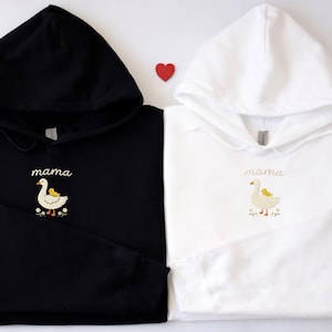 May include: A black hoodie and a white hoodie, each featuring an embroidered design of a white goose with a small yellow duckling on its back, accompanied by the word mama in cursive script and small flowers at the base of the embroidery.