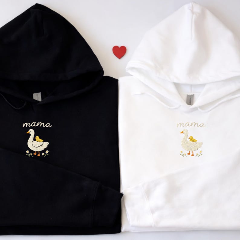 May include: A black hoodie and a white hoodie, each featuring an embroidered design of a white goose with a small yellow duckling on its back, accompanied by the word mama in cursive script and small flowers at the base of the embroidery.