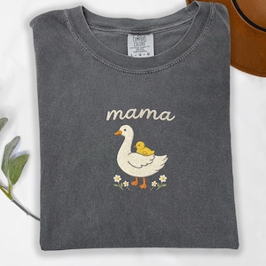 May include: A charcoal gray crewneck t-shirt featuring an embroidered design of a white goose with a small yellow duckling on its back. Above the goose, the word mama is embroidered in a cursive font. Small white flowers with yellow centers are below.