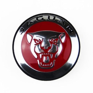 May include: A circular emblem featuring a metallic silver jaguar head with an open mouth, set against a vibrant red background. The word JAGUAR is embossed in silver capital letters along the upper curved border of the circular badge.