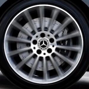 Può includere: Una ruota in lega argentata a più razze in movimento con uno pneumatico nero. Il coprimozzo centrale presenta il logo circolare Mercedes-Benz con la scritta Mercedes Benz. È visibile una pinza freno nera con il marchio Mercedes Benz.