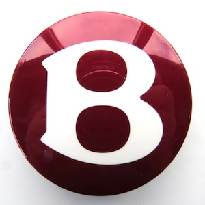 Può includere: Un emblema circolare color amaranto con finitura lucida. L'emblema presenta una grande lettera "B" bianca al centro. Il design è semplice e audace, con un netto contrasto tra la lettera bianca e lo sfondo amaranto.