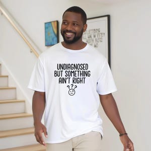 May include: White t-shirt with the text "UNDIAGNOSED BUT SOMETHING AIN'T RIGHT" in black, along with a confused face graphic. The shirt is worn by a person. The background includes a staircase and framed artwork.