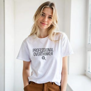 Pode incluir: Camiseta branca com o texto preto "PROFESSIONAL OVERTHINKER" e um desenho de lupa. A camiseta é de gola redonda. A modelo está usando calças marrons.