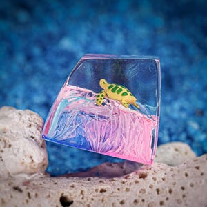May include: A translucent resin keycap featuring a miniature yellow and green sea turtle resting on a sculpted wave. The base of the keycap displays swirling patterns of pink, blue, and purple, creating a vibrant aquatic effect within the clear material.