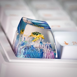 May include: A clear resin mechanical keyboard keycap featuring an encapsulated underwater scene with a yellow and green sea turtle swimming above blue and white coral-like structures. The keycap is positioned among standard white keyboard keys.
