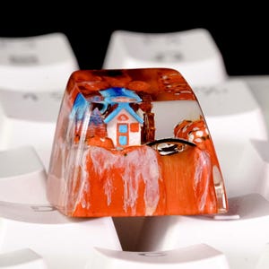 May include: A custom resin mechanical keyboard keycap featuring an embedded miniature scene of a small house with a blue roof and orange door, set against a vibrant orange and white landscape with textured details, resting on white keyboard keys.