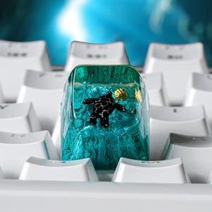 May include: A translucent teal resin mechanical keyboard keycap featuring an embedded black sea turtle figure and a small gold seashell accent. The keycap is positioned among several plain white keyboard keys on a standard computer keyboard.