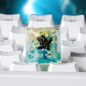May include: A clear resin mechanical keyboard keycap featuring an embedded black sea turtle figurine swimming amidst blue and white swirling water effects, positioned centrally among white keyboard keys on a computer keyboard.