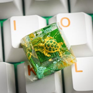 May include: A square, translucent resin mechanical keyboard keycap featuring an embedded yellow and green sea turtle figurine surrounded by green aquatic plants and clear water-like textures, positioned atop white keyboard keys.