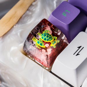 May include: A custom resin mechanical keyboard keycap featuring a deep red, translucent base with embedded miniature yellow and green sea turtles. The keycap is positioned between a purple keycap with green F1 text and a white keycap with black arrows.