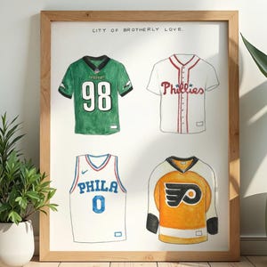 Könnte beinhalten: Gerahmter Aquarelldruck mit vier Sporttrikots. Die Trikots repräsentieren Teams aus Philadelphia: ein grünes Football-Trikot mit der Nummer 98, ein weißes Baseball-Trikot mit "Phillies", ein weißes Basketball-Trikot mit "PHILA 0" und ein orangefarbenes Hockey-Trikot.