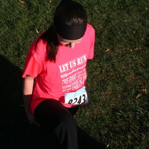 May include: A woman wearing a pink shirt with the text "Let Us Run With Endurance The Race That Is Set Before Us Keeping Our Eyes On Jesus" and a number "8243" on the front.