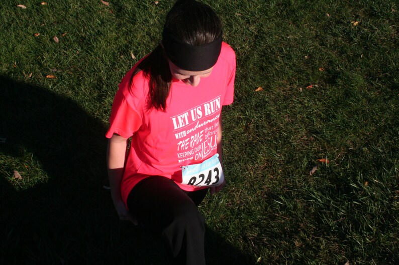 May include: A woman wearing a pink shirt with the text "Let Us Run With Endurance The Race That Is Set Before Us Keeping Our Eyes On Jesus" and a number "8243" on the front.