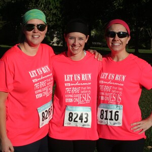May include: Three women wearing pink shirts with the text "Let Us Run With Endurance The Race That Lies Before Us Keeping Our Eyes On Jesus" and race numbers. The women are smiling and standing together.