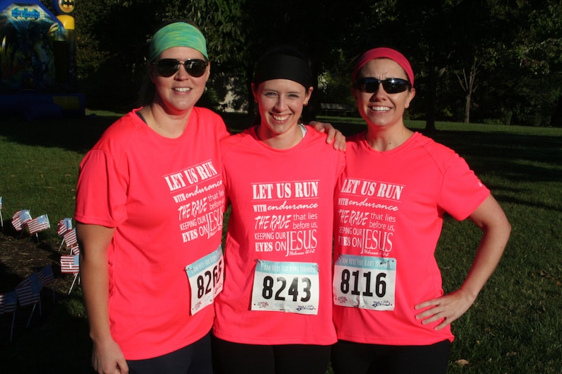 May include: Three women wearing pink shirts with the text "Let Us Run With Endurance The Race That Lies Before Us Keeping Our Eyes On Jesus" and race numbers. The women are smiling and standing together.
