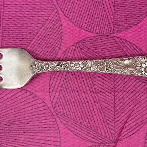 May include: A silver fork with four prongs and a floral design on the handle. The fork is placed on a pink background with a geometric pattern.