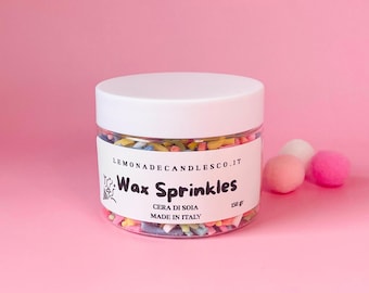 May include: A clear jar with a white lid filled with colorful wax sprinkles. The label reads "Wax Sprinkles" and "Cera di Soia Made in Italy". The jar is next to three small, round, pink and white pom-poms. The background is a solid pink.