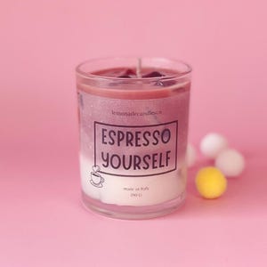 Op de afbeelding: Een gelaagde glazen kaars met een witte basis, een paarsroze midden en een donkerroze bovenkant. Het etiket luidt "ESPRESSO YOURSELF" met een koffiekopje. Gemaakt in Italië, 190 g. Roze achtergrond.