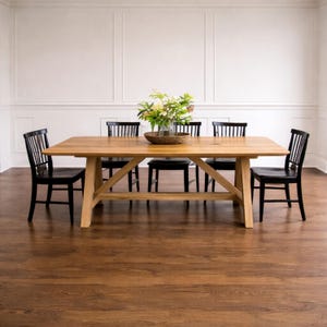 Op de afbeelding: Een licht houten eettafel met een schraag, omringd door vier zwarte stoelen. Een gevlochten mand met bloemen staat in het midden van de tafel. De kamer heeft witte muren en een donkere houten vloer.