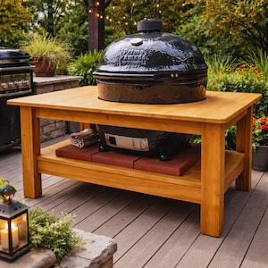 May include: A black Primo ceramic grill sits on a light brown wooden table with a lower shelf. The grill has a shiny, rounded lid and the table is outdoors on a wooden deck. The table is made of light brown wood.