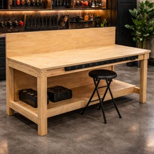 Puede incluir: Un banco de trabajo grande de madera clara con una regleta negra a lo largo del borde frontal. Una banqueta plegable negra está frente al banco de trabajo. Dos contenedores de almacenamiento negros están en el estante inferior. El banco de trabajo está en un taller.