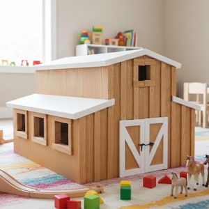 May include: A wooden toy barn with a white roof and trim, featuring a small window and double doors. The barn is made of light-colored wood and sits on a colorful rug. Toy horses and blocks are nearby.